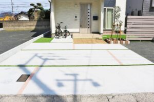 目地に人工芝を通した白いコンクリート舗装の駐車スペースと、タイル敷きのアプローチ