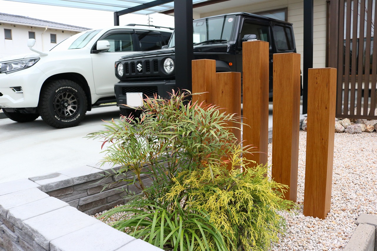 木製の装飾ポストと植栽が配置されたカーポート脇の駐車スペース、白と黒の2台の車