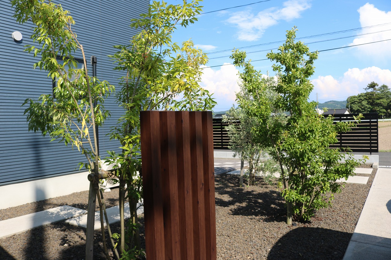 木調の門柱の周りに若木を植えた庭のシンボルツリー