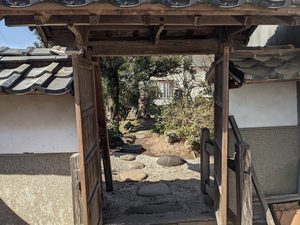 瓦屋根をのせた木製の門と、その先に続く飛び石と砂利敷きの通路
